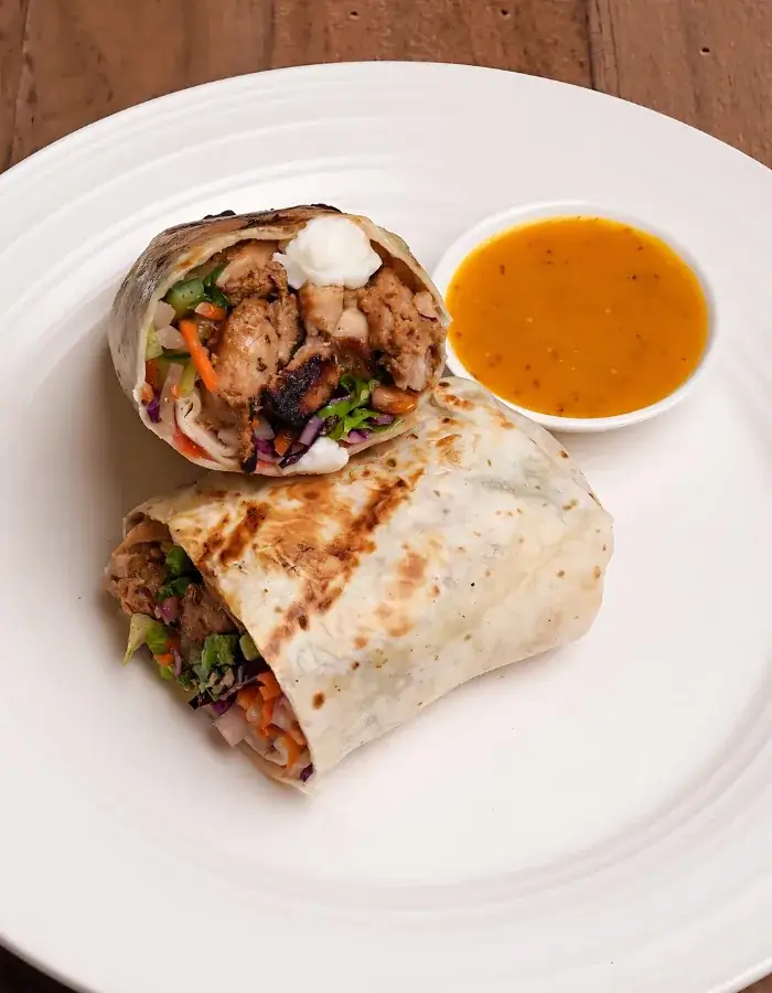 A grilled chicken wrap cut in half with a cup of sauce on a white plate